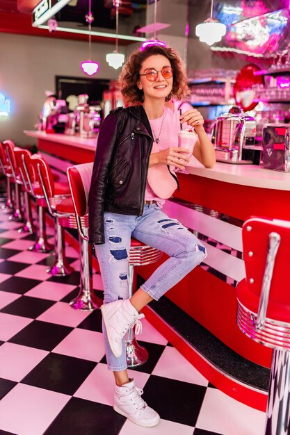 Hermosa mujer sonriente con estilo en un café retro de los años 50 sentado en el bar bebiendo un cóctel de batido de leche con camiseta rosa, chaqueta de cuero balcj y jeans divirtiéndose de buen humor