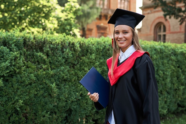 Hermosa mujer sonriente graduada en bata de graduación en campus universitario