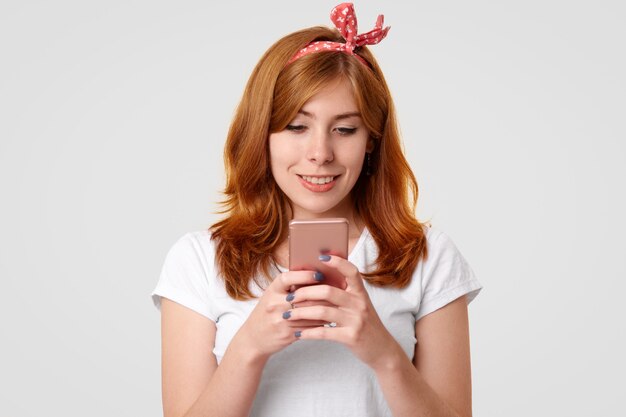 Hermosa mujer sonriente de jengibre con una sonrisa encantadora, utiliza un teléfono móvil moderno, mensajes con amigos