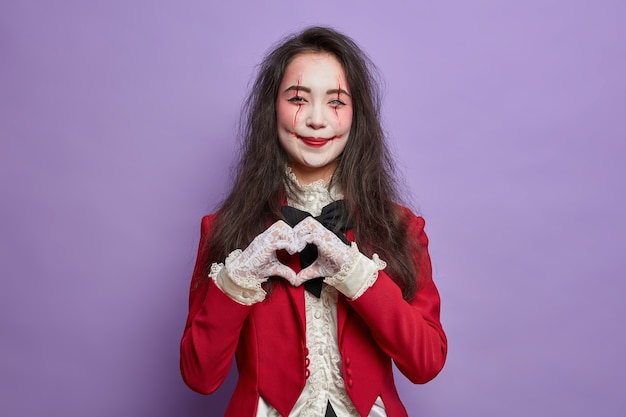 Hermosa mujer sonriente con maquillaje temeroso rostro de fantasma pálido y cicatrices sangrientas hace un gesto de corazón y expresa amor en la fiesta de Halloween aislada sobre una pared púrpura