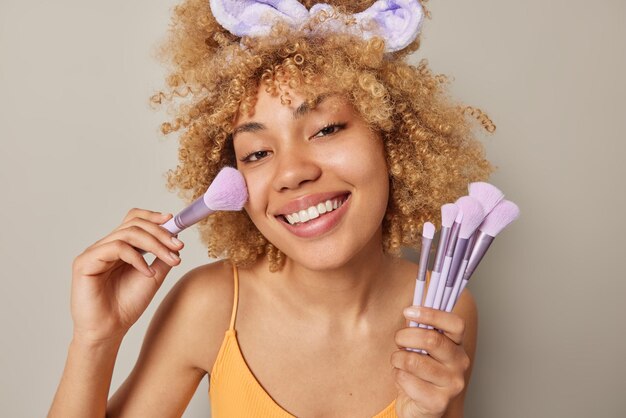 Hermosa mujer sonriente con el pelo rizado aplica rubor en la cara con cepillo cosmético en polvo mejillas mira felizmente a la cámara usa camiseta y diadema aislado sobre fondo gris Maquillaje diario