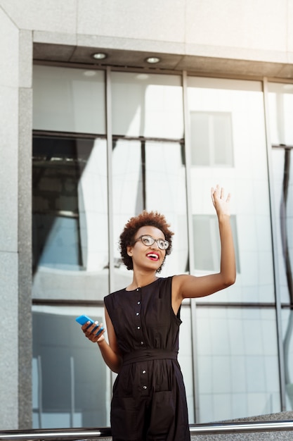 Foto gratuita hermosa mujer sonriente saludo sosteniendo teléfono caminando por la ciudad