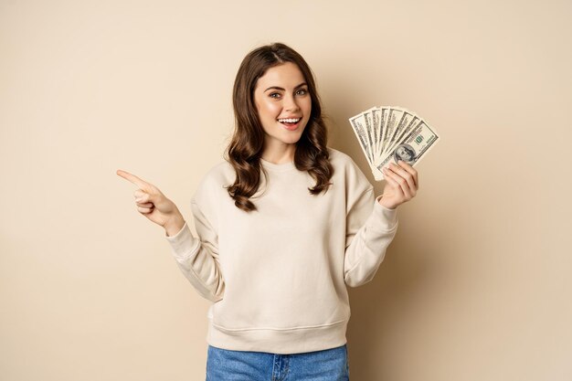 Hermosa mujer sonriente sosteniendo dólares de dinero y señalando con el dedo a la izquierda, mostrando la pancarta de la empresa con el logotipo, de pie sobre un fondo beige.