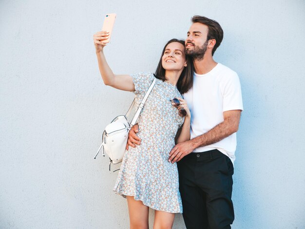 Hermosa mujer sonriente y su guapo novio Mujer con ropa informal de verano Familia feliz y alegre Mujer divirtiéndose Pareja posando en la calle cerca de la pared Abrazándose unos a otros Tomando selfie