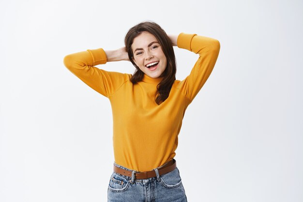 Hermosa mujer sonriente tomados de la mano detrás de la cabeza y sonriendo despreocupado, descansando en el ocio, de pie contra la pared blanca