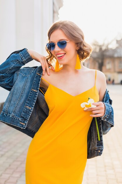 Hermosa mujer sonriente en vestido amarillo elegante con chaqueta de mezclilla, atuendo de moda, tendencia de moda primavera verano, estado de ánimo soleado, feliz, gafas de sol azules, moda callejera