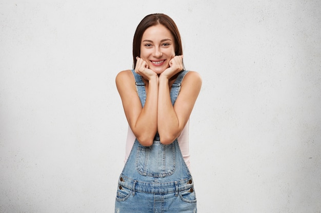 Hermosa mujer con sonrisa encantadora con expresión alegre en su rostro, tomados de la mano en su rostro, satisfecha con un cumplido o un bonito regalo.