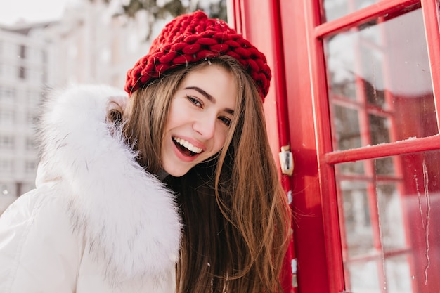 Hermosa mujer con sonrisa feliz posando cerca de la cabina de teléfono roja en la mañana de diciembre. Retrato al aire libre de la maravillosa dama europea viste gorro de punto y bata blanca en invierno.