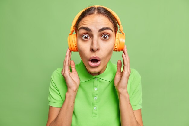 Hermosa mujer sorprendida con apariencia europea escucha música o audo prodcast a través de auriculares inalámbricos vestida de manera informal, no puede creer en noticias increíbles aisladas sobre una pared verde