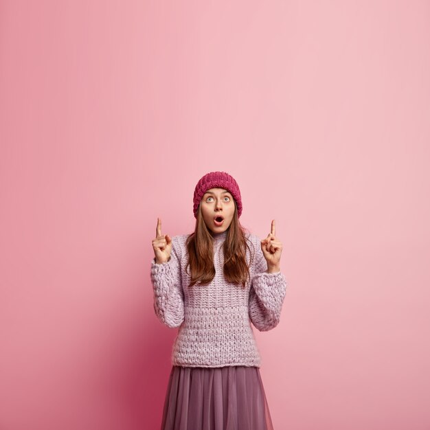 Hermosa mujer sorprendida con la boca muy abierta, vestida con sombrero, jersey de punto y falda larga, puntos arriba en el espacio en blanco para su contenido publicitario. Gente, asombro, publicidad