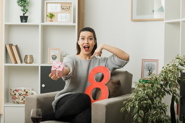 Hermosa mujer sorprendida en el día de la mujer feliz sosteniendo y apunta actualmente sentado en un sillón en la sala de estar