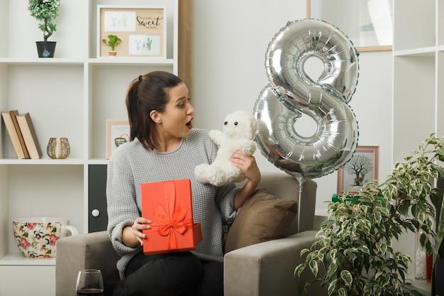 Hermosa mujer sorprendida en el día de la mujer feliz sosteniendo el presente y mirando el oso de peluche en su mano sentado en un sillón en la sala de estar