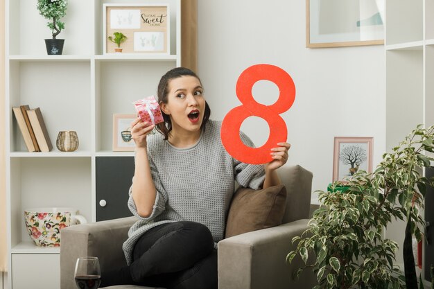 Hermosa mujer sorprendida en el día de la mujer feliz sosteniendo presente con el número ocho sentado en un sillón en la sala de estar