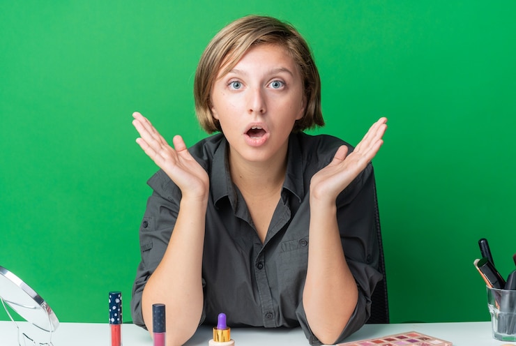 Foto gratuito hermosa mujer sorprendida se sienta a la mesa con herramientas de maquillaje extendiendo las manos