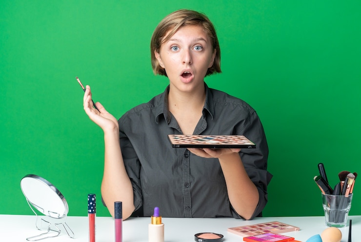 Foto gratuito hermosa mujer sorprendida se sienta a la mesa con herramientas de maquillaje con paleta de sombra de ojos con pincel de maquillaje