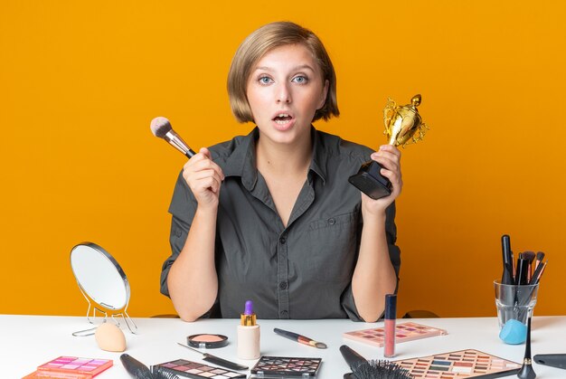 Hermosa mujer sorprendida se sienta a la mesa con herramientas de maquillaje sosteniendo la taza ganadora con pincel de maquillaje