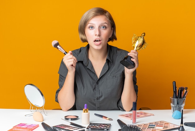 Hermosa mujer sorprendida se sienta a la mesa con herramientas de maquillaje sosteniendo la taza ganadora con pincel de maquillaje