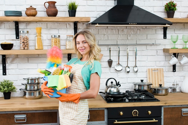 Hermosa mujer sosteniendo el cubo de herramientas y productos de limpieza en la cocina