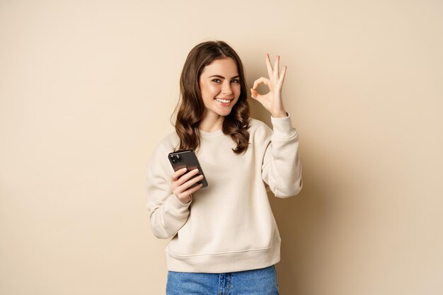 Hermosa mujer sosteniendo un teléfono celular, un teléfono móvil y un cartel bien, recomendando una aplicación, una aplicación de compras, de pie sobre un fondo beige.