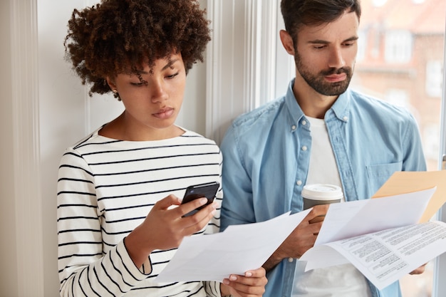 Hermosa mujer en suéter a rayas hace contabilidad en el teléfono celular, verifica las cifras en el documento