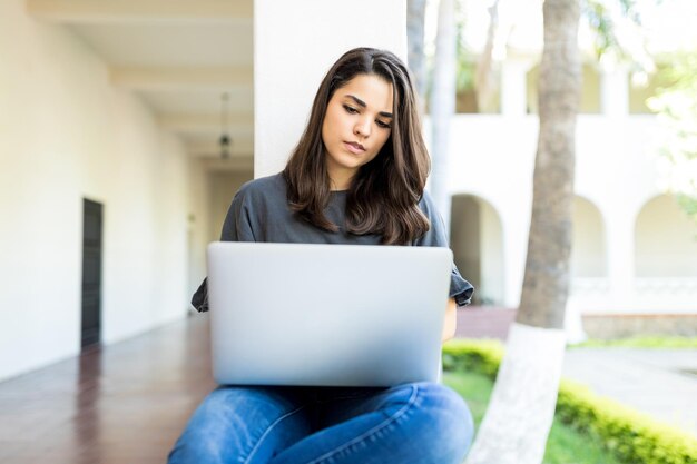 Hermosa mujer tecleando en la computadora portátil mientras está sentada en la baranda en el campus