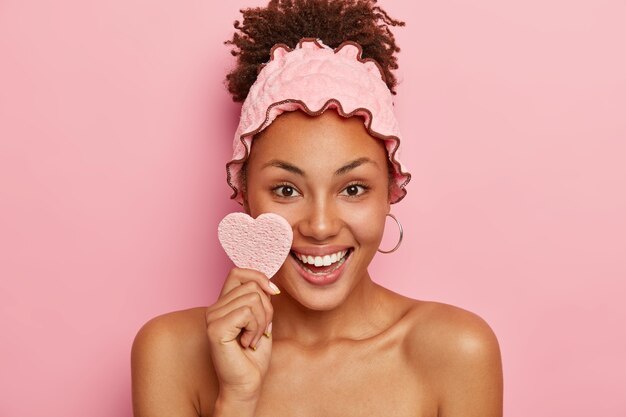 Hermosa mujer tiene una piel sana, se limpia la cara con una esponja cosmética, sonríe positivamente, muestra los dientes blancos, usa una diadema para la ducha, se ducha después del trabajo, aislada en la pared rosa