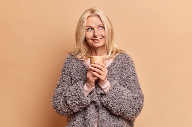 Hermosa mujer tierna ha complacido la expresión pensativa mira hacia otro lado sueños sobre poses de descanso con una taza de café de papel disfruta bebiendo bebidas aromáticas contra la pared beige