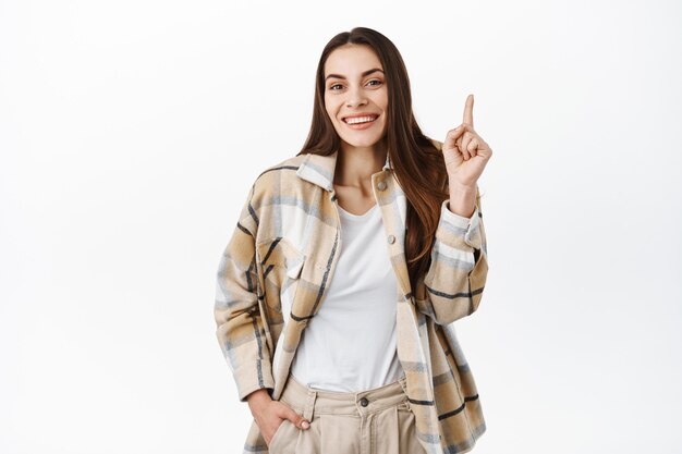 Hermosa mujer tímida apuntando con el dedo hacia arriba y riendo, sonriendo lindo, mostrando publicidad, mostrando el banner del producto o el logotipo de la empresa en el espacio de copia superior, de pie sobre una pared blanca