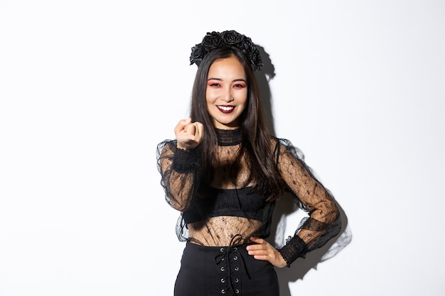 Hermosa mujer en traje de bruja diciendo que se acerque, mueva el dedo y sonriendo, de pie con traje de halloween, maquillaje gótico, de pie sobre fondo blanco.