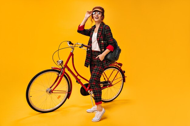 Hermosa mujer en traje a cuadros y gorra posando con bicicleta Chica de moda en anteojos y traje moderno mirando a la cámara