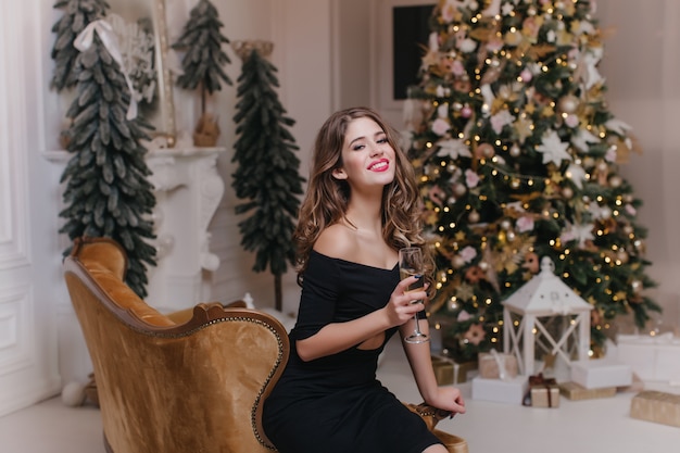Hermosa mujer en traje negro sentada en un sofá marrón con una copa de vino. Retrato de niña confiada de pelo largo celebrando las vacaciones de invierno con árbol de Navidad en la pared.