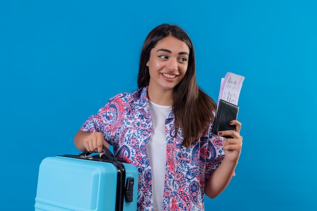 Foto gratuita hermosa mujer turista con maleta de viaje y pasaporte con boletos mirando a un lado con una sonrisa en la cara concepto de viaje feliz y positivo de pie sobre el espacio azul