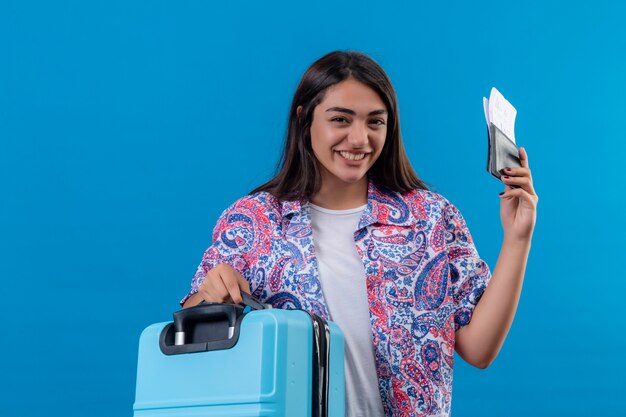 Hermosa mujer turista con maleta de viaje y pasaporte con boletos con sonrisa en la cara feliz y positivo concepto de viaje de pie sobre el espacio azul
