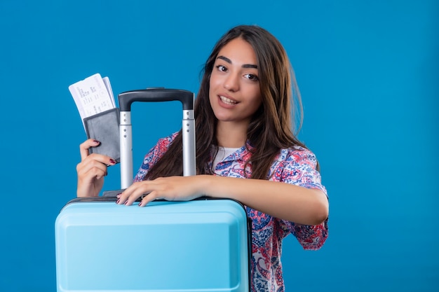 Hermosa mujer turista con maleta de viaje y pasaporte con boletos con sonrisa en la cara feliz y positivo concepto de viaje de pie sobre el espacio azul