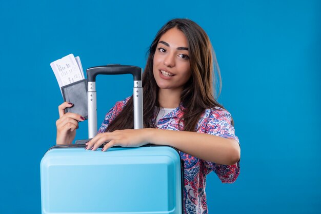 Hermosa mujer turista con maleta de viaje y pasaporte con boletos con sonrisa en la cara feliz y positivo concepto de viaje de pie sobre el espacio azul