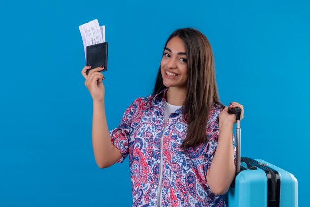 Hermosa mujer turista con maleta de viaje y pasaporte con boletos con sonrisa en la cara feliz y positivo concepto de viaje de pie sobre el espacio azul