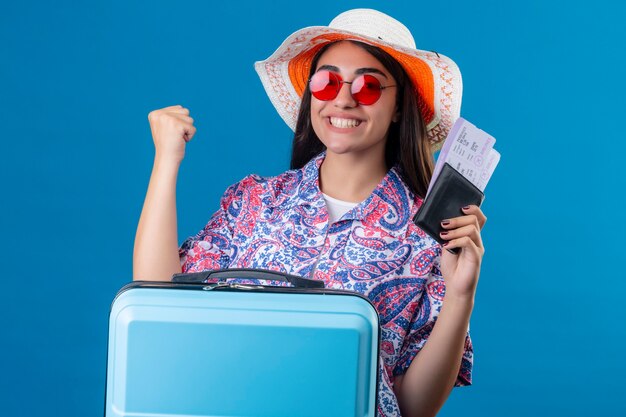 Hermosa mujer turista con sombrero de verano con gafas de sol rojas sosteniendo la maleta de viaje y pasaporte con boletos con una sonrisa en la cara feliz y positiva levantando el puño después de una victo