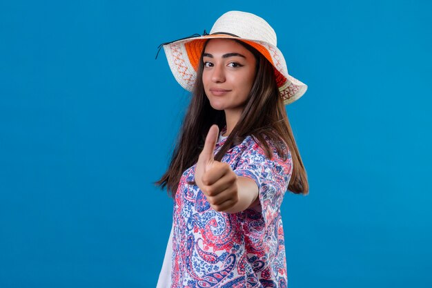 Hermosa mujer turista con sombrero de verano positivo y feliz sonriendo mostrando los pulgares para arriba sobre el espacio azul