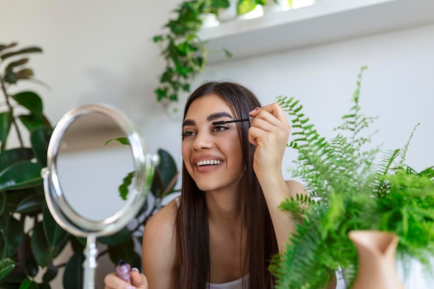 Hermosa mujer usando rímel en las pestañas en el baño en el tiempo de la mañana Sonriente mujer joven aplicando maquillaje de ojos y mirando al espejo Chica de belleza aplicando rímel negro en el baño en casa