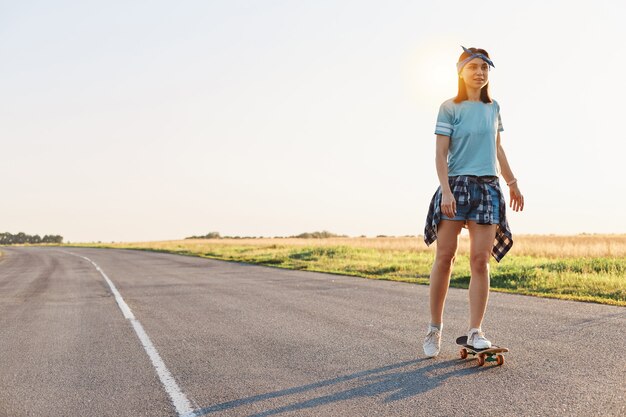 Hermosa mujer vestida con camiseta, banda corta y el pelo en patineta en la calle, mirando en la distancia, pasando tiempo a solas con placer, estilo de vida activo y saludable.