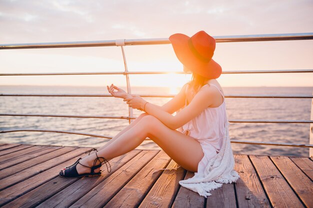 Hermosa mujer en vestido blanco sentada junto al mar al amanecer en estado de ánimo romántico con sombrero rojo