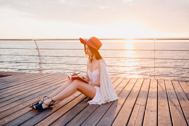 Hermosa mujer en vestido blanco sentada junto al mar al amanecer pensando y tomando notas