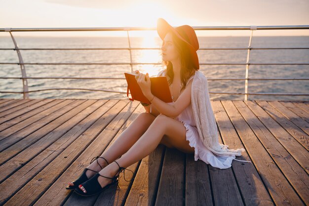 Hermosa mujer en vestido blanco sentada junto al mar en el amanecer pensando y haciendo notas en el libro diario