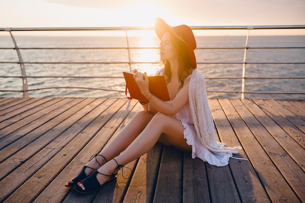 Hermosa mujer en vestido blanco sentada junto al mar en el amanecer pensando y haciendo notas en el libro diario