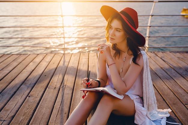 Hermosa mujer en vestido blanco sentada junto al mar en el amanecer pensando y haciendo notas en el libro diario