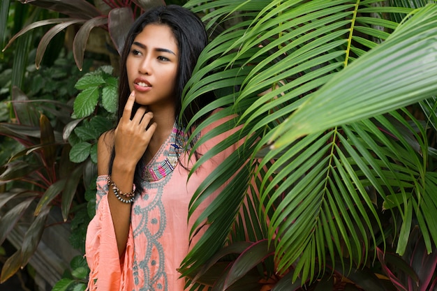 Hermosa mujer en vestido boho con hojas de palmera verde. Hermosa mujer asiática en ropa de verano de moda y accesorios posando en retrato de naturaleza tropical.