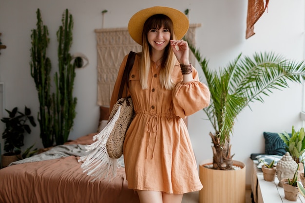 Hermosa mujer en vestido de lino y sombrero de paja posando en apartamento de estilo boho