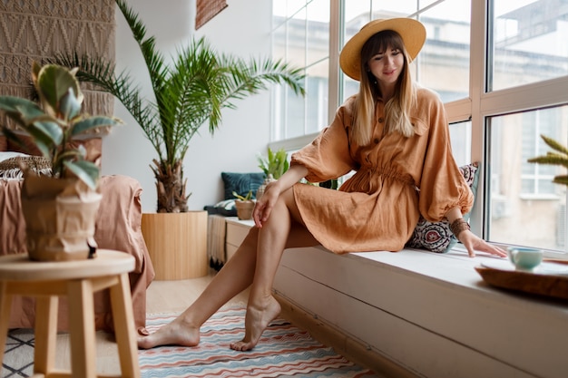 Hermosa mujer en vestido de lino y sombrero de paja posando en apartamento de estilo boho