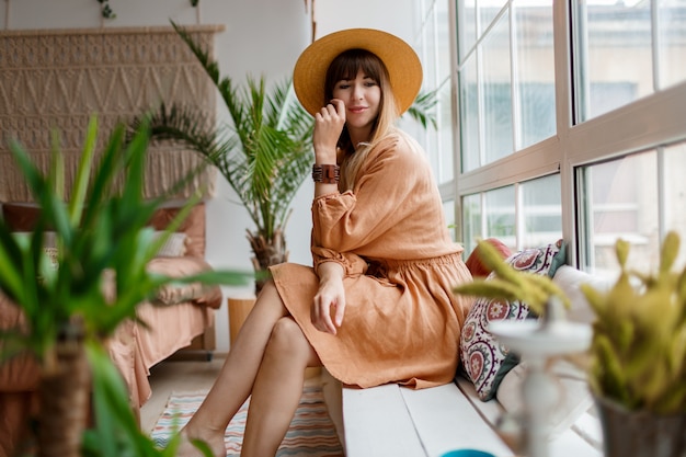 Hermosa mujer en vestido de lino y sombrero de paja posando en apartamento de estilo boho