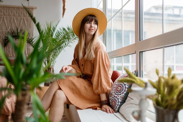 Hermosa mujer en vestido de lino y sombrero de paja posando en apartamento de estilo boho